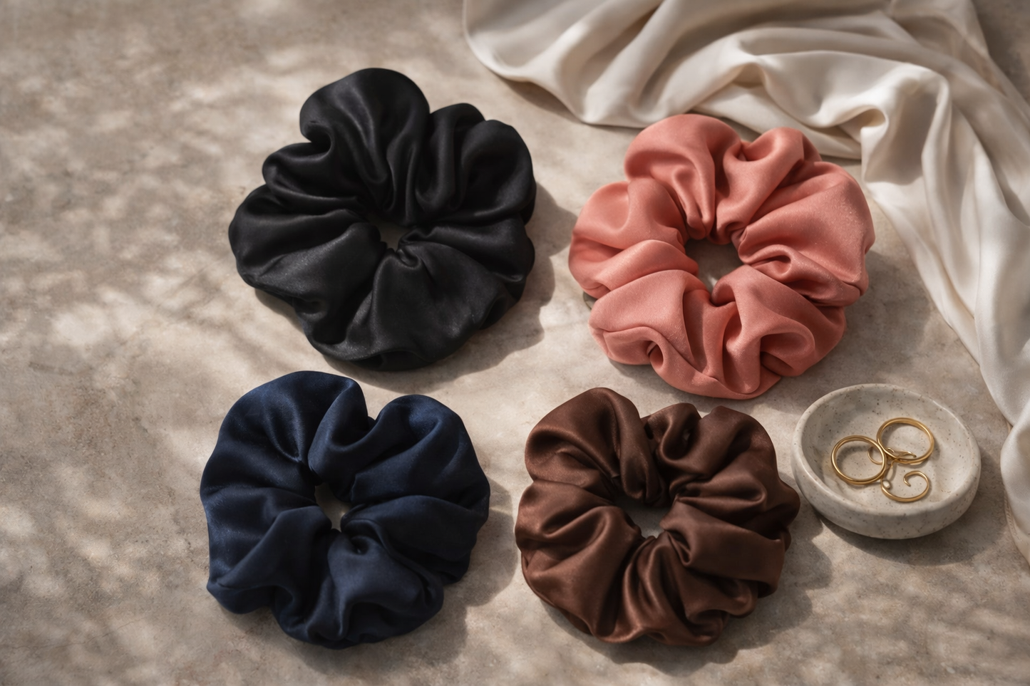 ASORA Silk Scrunchie, Four scrunchies in black, pink, blue, and brown on a textured surface with a small dish containing gold rings.