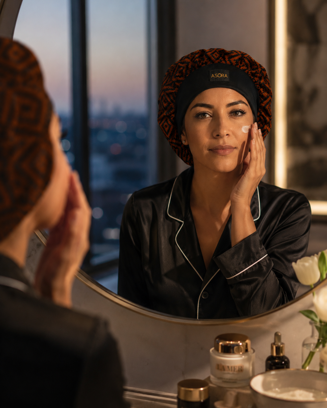 Woman wearing ASORA crown bonnet applying cream to her face in front of a mirror with a cityscape view.
