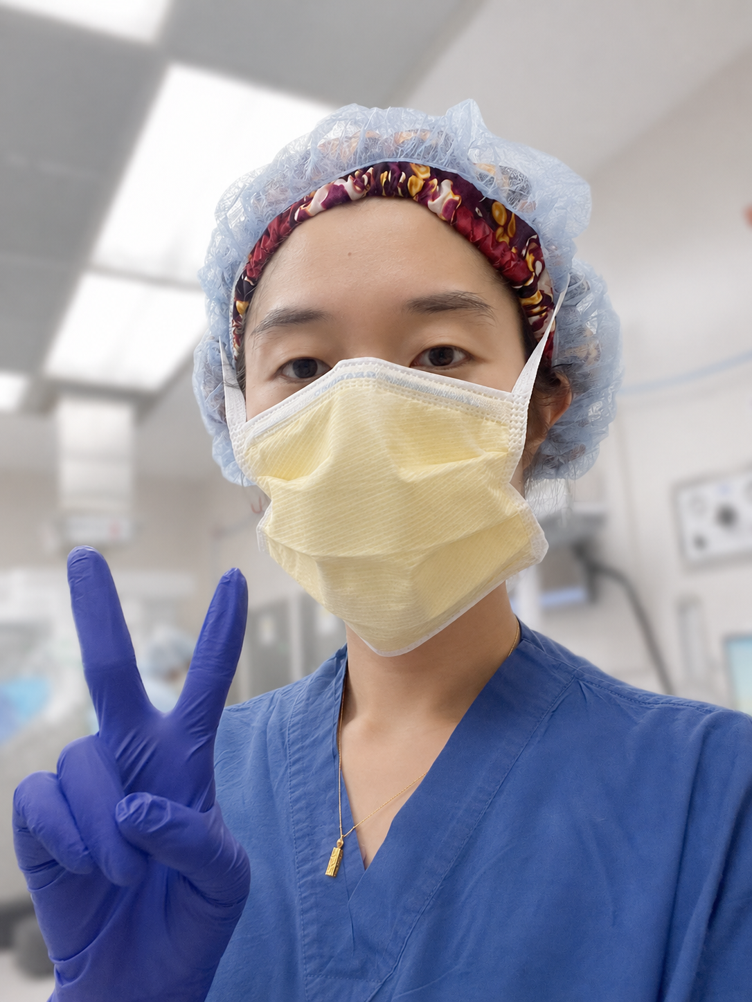 Person in medical scrubs, ASORA dara bonnet, and gloves making a peace sign in a hospital setting