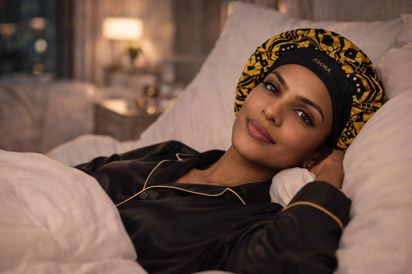 Woman wearing a black and yellow patterned ASORA Crown Bonnet, lying in bed in a cozy bedroom.